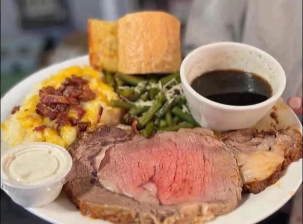 A thick slice of prime rib with loaded mashed potatoes, green beans, a dinner roll and a cup of au jus.